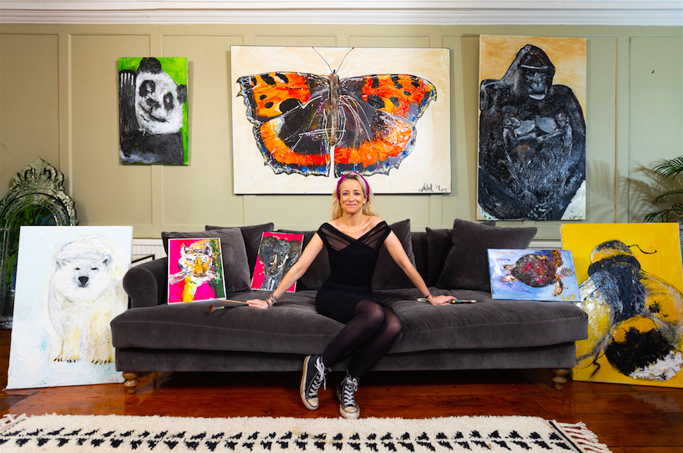 Artist Leah Wood sits on a black sofa surrounded by a collection of her art work from left to right a Polar Bear, a panda, a tiger, an elephant, a butterfly, a gorilla, a turtle and a bumble bee.